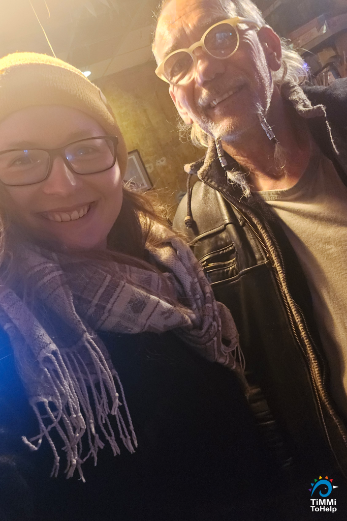 Das Foto ist ein Selfie und zeigt zwei Personen, die nebeneinander stehen und in die Kamera lächeln. Links ist eine Frau, es ist Saskia. Sie trägt eine Brille mit einem dunklen Rahmen, eine gelbe Mütze und einen gemusterten Schal.
Rechts ist ein Mann, es ist Armin. Er hat eine helle, runde Brille, hat graue Haare und einen Bart, der in zwei kleinen Zöpfen geflochten ist. Er trägt eine schwarze Lederjacke über einem hellen Oberteil.